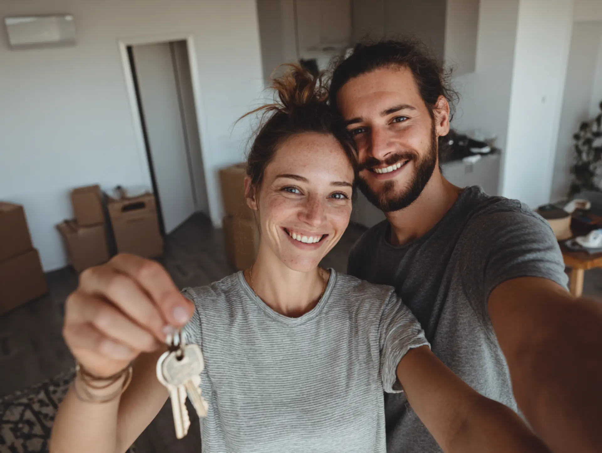 Pareja joven sonriendo y mostrando las llaves de su nuevo piso con cajas de mudanza al fondo.
