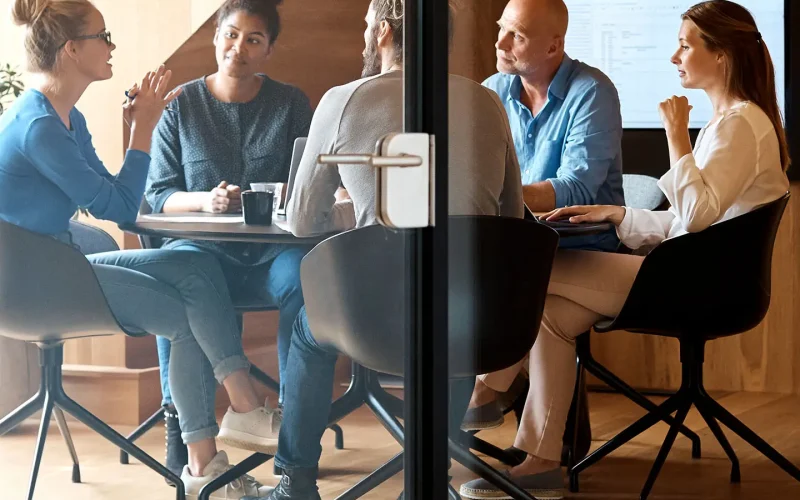 Equipo de cuatro personas reunido alrededor de una mesa en una sala acristalada con pantalla de presentación.