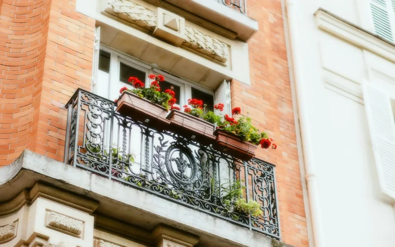 Balcón con barandilla de hierro forjado y flores rojas en un edificio de ladrillo visto desde la calle.