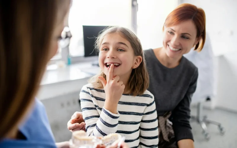 Niña señalando sus dientes ante la dentista mientras su madre sonríe a su lado.