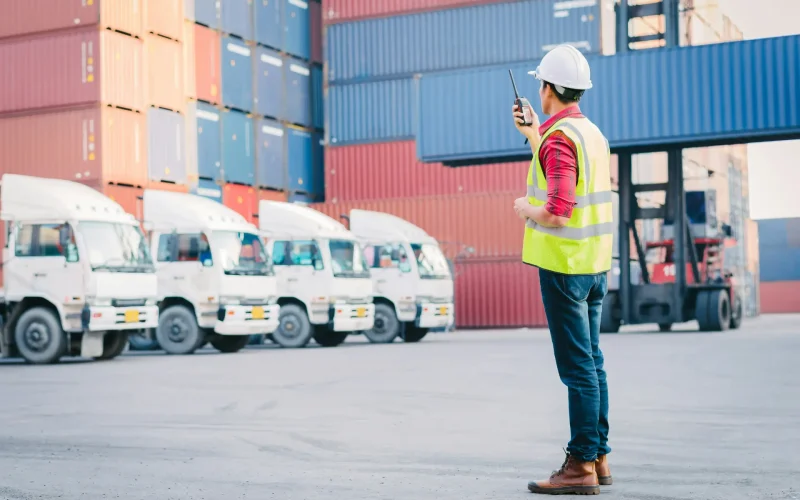Supervisor con chaleco y casco coordinando camiones y contenedores en un puerto de carga.