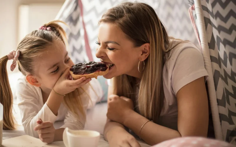 Madre e hija jugando y compartiendo un donut dentro de un tipi de tela en casa.