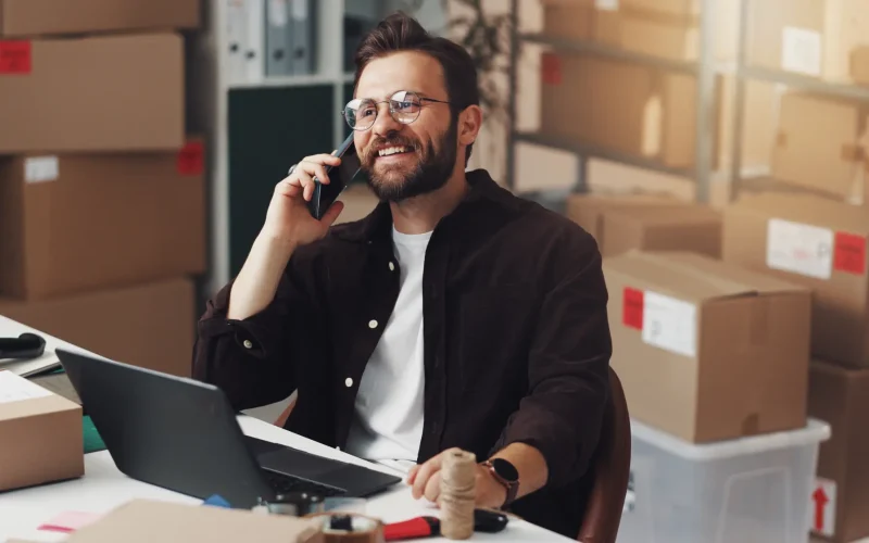 Autónomo sonriente hablando por teléfono en su almacén rodeado de cajas de cartón y portátil.